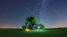 man with a flashlight next to a tent and tree underneath a meteor shower