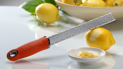 A microplane premium grater zester with red handle with lemon peel and whole lemons in bowl in background
