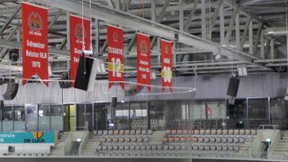 Silver Alcons Audio speakers hang from the rafters in front of several red banners at an ice hockey arena.