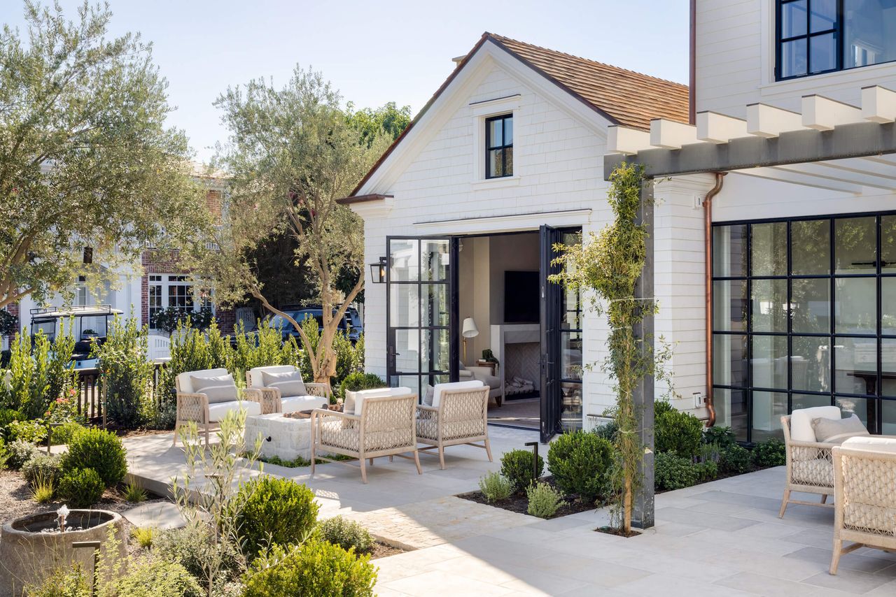 a sunny patio area with furniture, evergreen plants and a white house