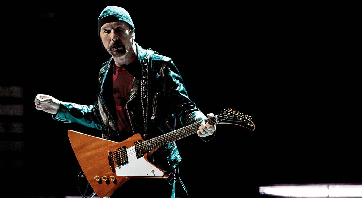 The Edge of U2 plays his 1976 Gibson Explorer onstage in Milan, 2018. He wears a beanie and is illuminated by a white stagelight. The Explorer has a natural finish and a white pickguard.