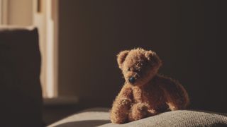 Photo of a teddy bear sitting on a bed in a darkened room with light coming through a window