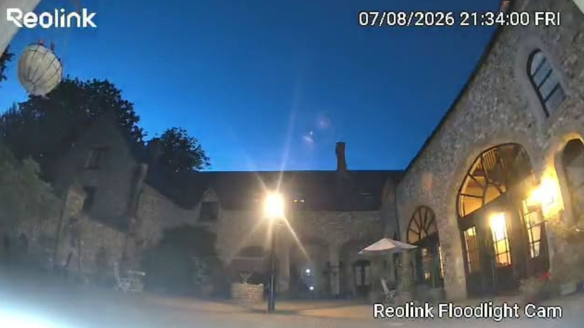 Reolink camera night footage of stone courtyard building, floodlight illuminating archways and doorway.