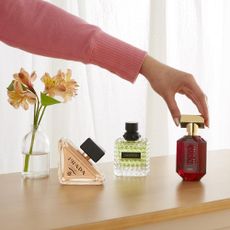 Fragrances on a shelf with a flower in a vase and arm reaching out touching the 3rd bottle