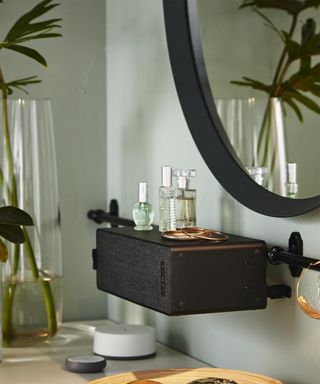 A bedroom including Ikea's SYMFONISK WiFi bookshelf speaker, pastel green wall decor, black-framed mirror with perfumes and colognes