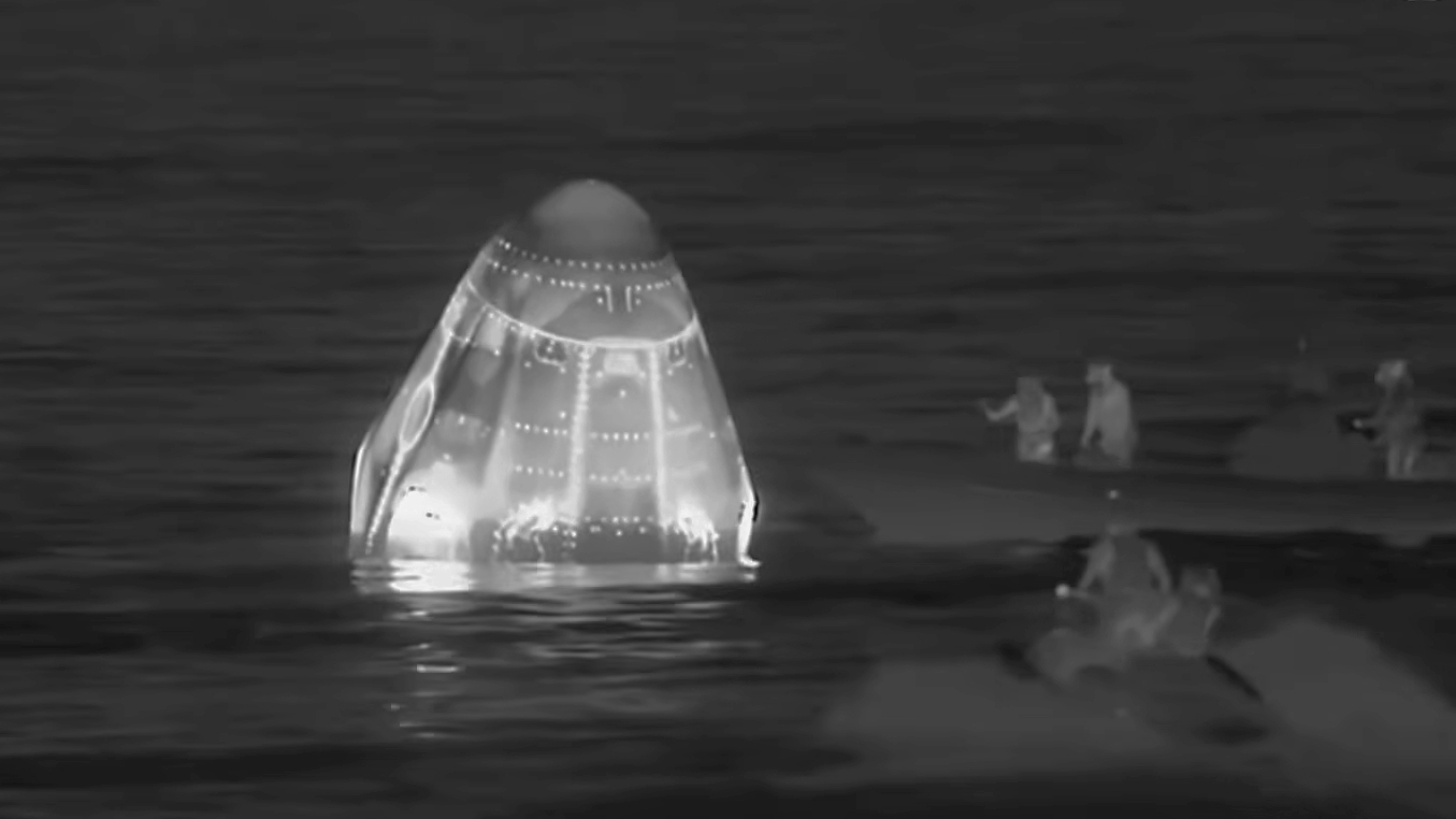 infrared black-and-white image of a conical white space capsule floating in the ocean, with a handful of people in small boats approaching it