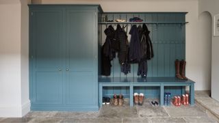 Traditional boot room with stone flagstone flooring, a built-in teal storage unit with coat hooks, bench seating, and cubby holes for shoes. Various outerwear and boots are neatly arranged, creating a functional and organized space.