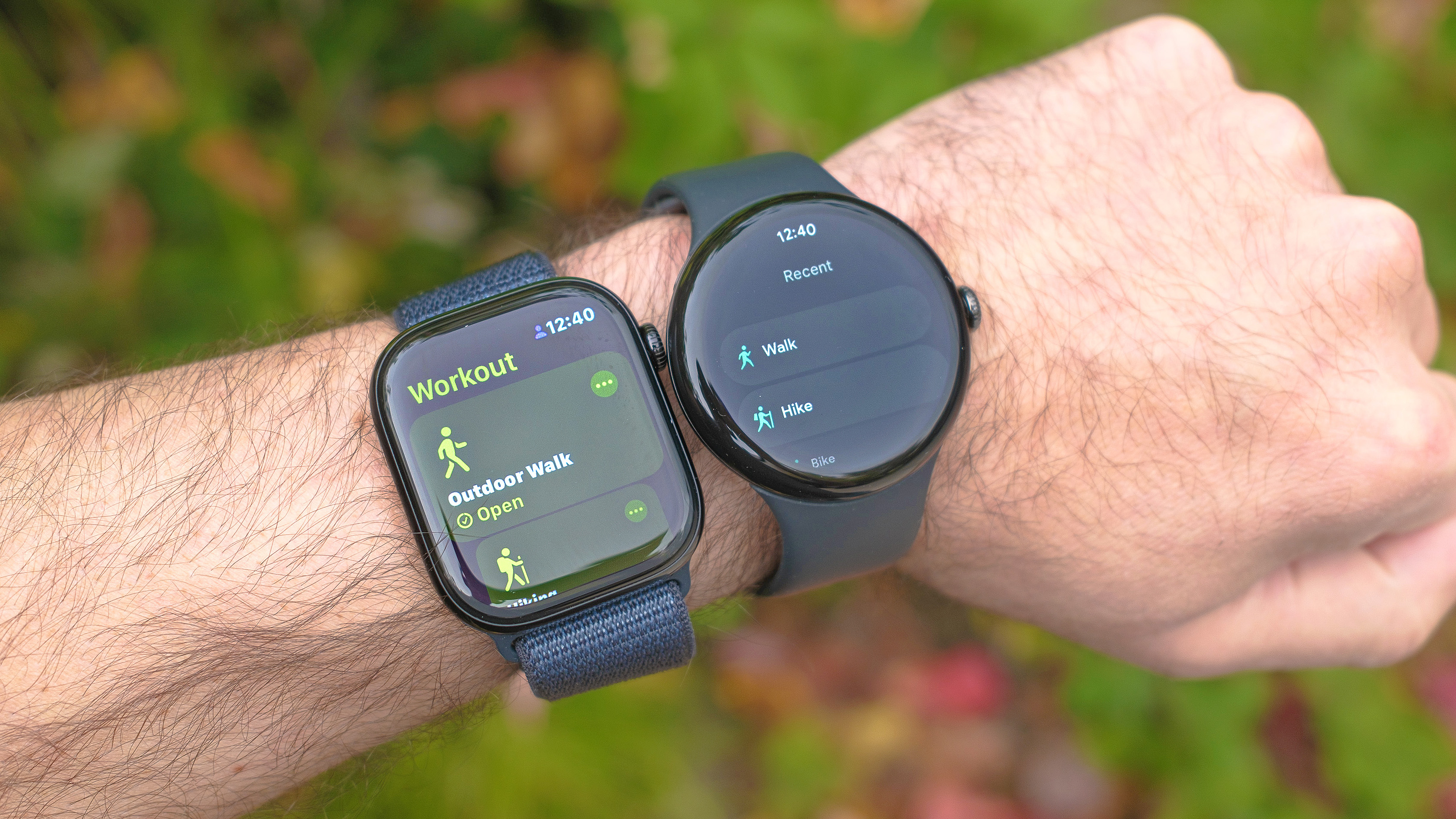 Close-up of Apple Watch 10 (left) next to Pixel Watch 3 on a user's wrist. In the background are colorful leaves that are out of focus.