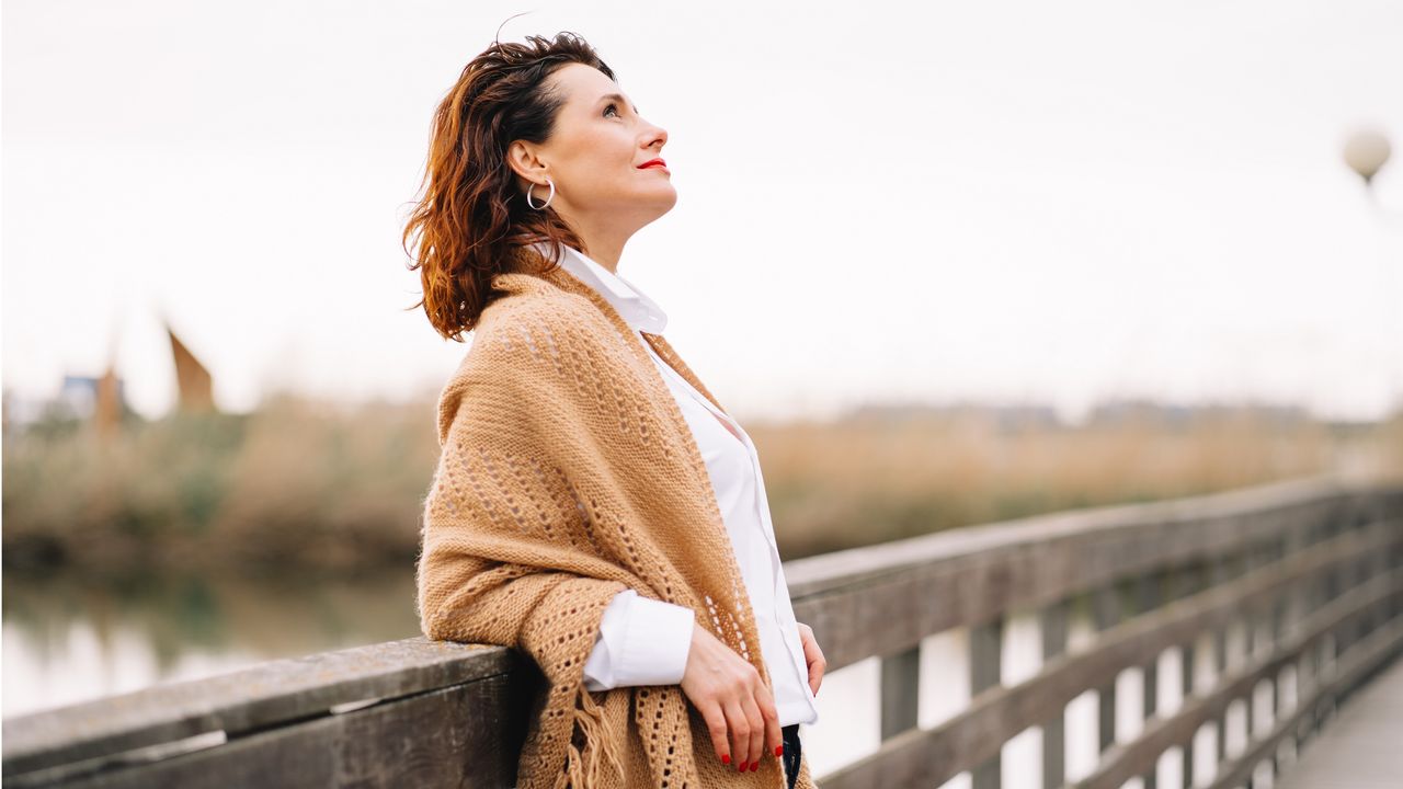 A woman leans back against the railing of a footbridge and looks thoughtfully toward the sky. 