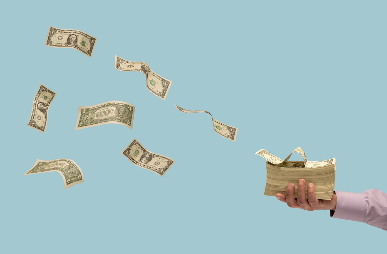 Man&amp;#039;s hand holding stack of US currency with some bills flying away, blue background