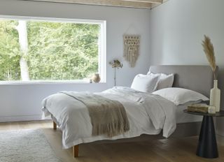 white bedroom/apartment bedroom with upholstered bed, modern black side table, wall hanging, wooden floorboards, rug, oatmeal colored blanket, white bedding