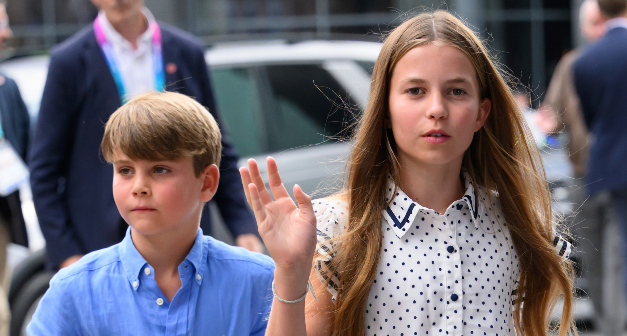Princess Charlotte waving in a white dress walking in front of Prince Louis