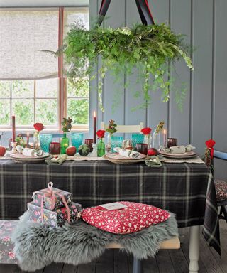 a dining area with a black tartan tablecloth, a bench set with a grey sheepskin rug and presents on top, red and green themed table dressings, wood panel walls and a chandelier made of real foliage hanging over the table