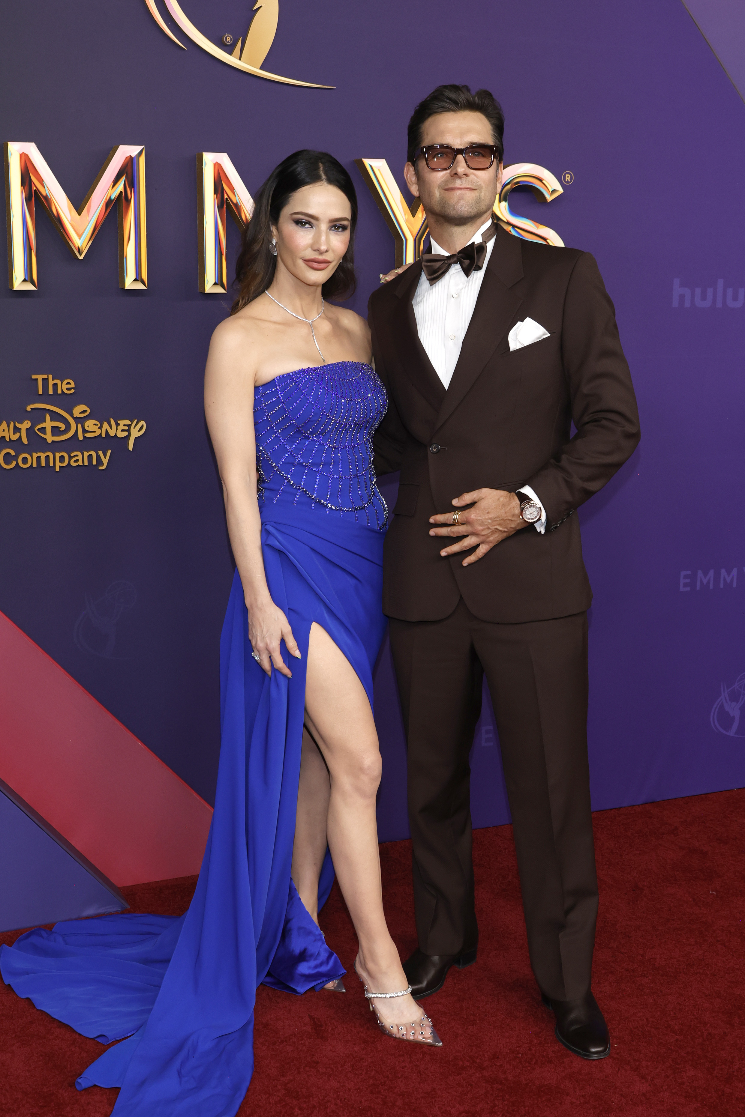 Sandra Vergara wears a blue dress and Antony Starr wears a brown suit as they stand on the red carpet at the 76th Primetime Emmy Awards at Peacock Theater on September 15, 2024 in Los Angeles, California