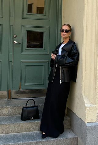 a black maxi skirt outfit shown on a woman wearing black sunglasses with a black leather jacket layered over a white t-shirt with a black maxi pencil skirt with pointed pumps and a black handbag
