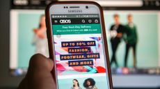 Asos website homepage pictured on a smartphone screen and in desktop monitor in background, with smartphone held in hand in foreground.
