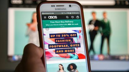 Asos website homepage pictured on a smartphone screen and in desktop monitor in background, with smartphone held in hand in foreground.