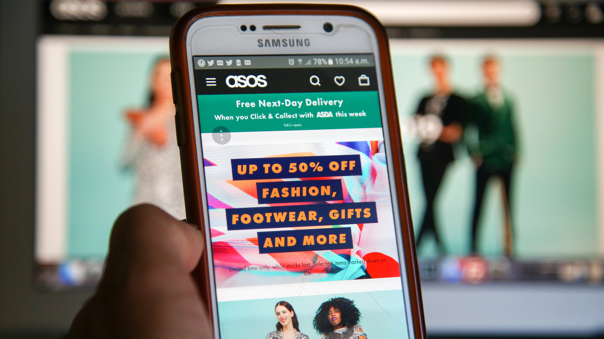 Asos website homepage pictured on a smartphone screen and in desktop monitor in background, with smartphone held in hand in foreground.