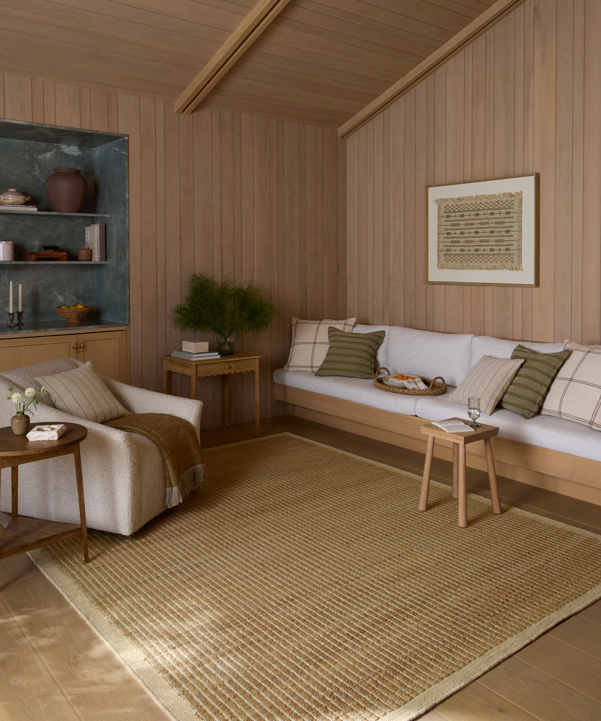 a warm living room with wood paneled walls and ceiling, a built-in couch with white cushions, an accent chair, and a jute rug