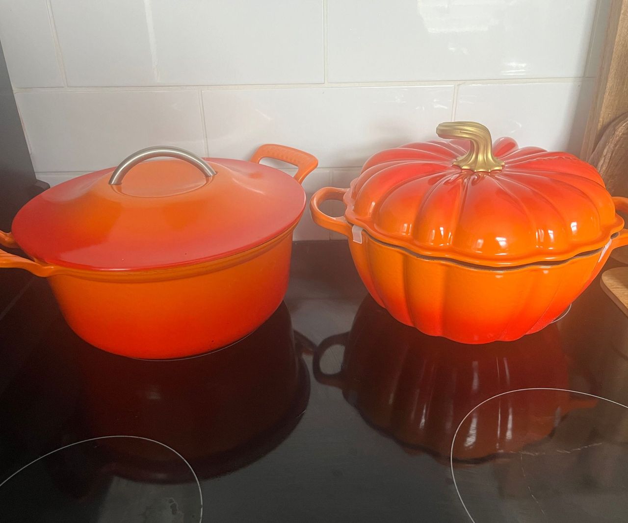 An orange Le Creuset Pumpkin cocotte on a black stove, next to an orange Le Creuset modern heritage Dutch oven.