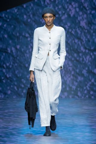 A female model wearing a white long sleeved button up shirt, white pants and black sandals walking down a runway.