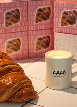 A large croissant beside a mug candle on a tiled surface with colorful packaging in the background