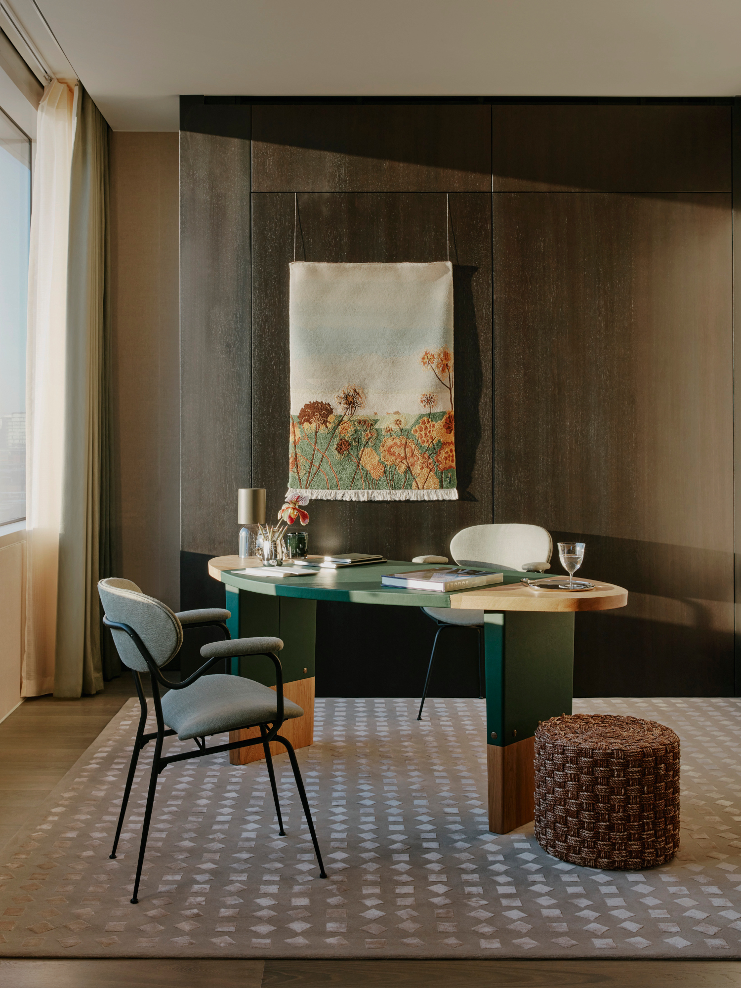 Home office with dark wood clad walls, green oval desk, blue chair, floral tapestry and beige geometric rug