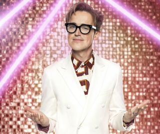 Tom Fletcher sports a white suit over a patterned shirt as he stands in front of a golden glittery backdrop