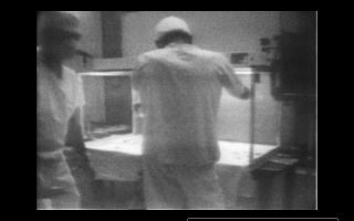 A still image taken from a digitally transcribed version of the original 16mm film of the microbial sampling of the camera. Two investigators stand in front of the laminar flow bench in which the camera resided during sampling. Note that they are wearing masks and head coverings that do not fully cover their heads, short-sleeve scrubs that do not extend below the level of the bench-top (allowing air potentially to blow into and out of them), and gloves over their hands but with no protection for their arms.