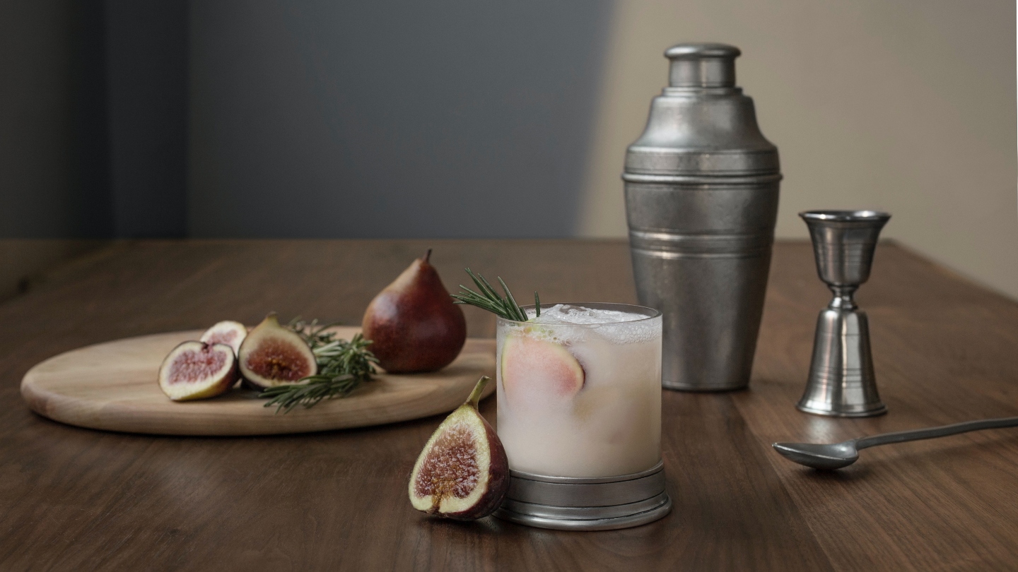 Silver cocktail shaker and shot glass behind a chopping board with figs on it and a glass with a cocktail in it