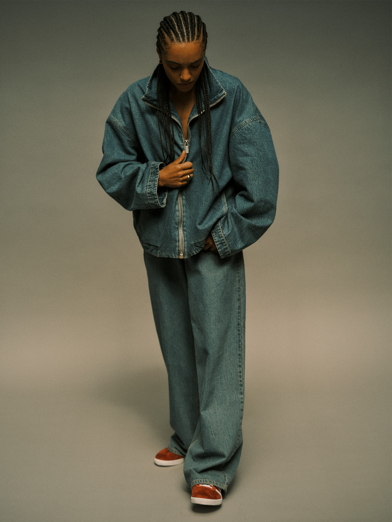 Actor Maisie Richardson-Sellers poses on a gray seamless backdrop wearing an oversize zip-up denim jacket and baggy jeans with red Adidas sneakers.