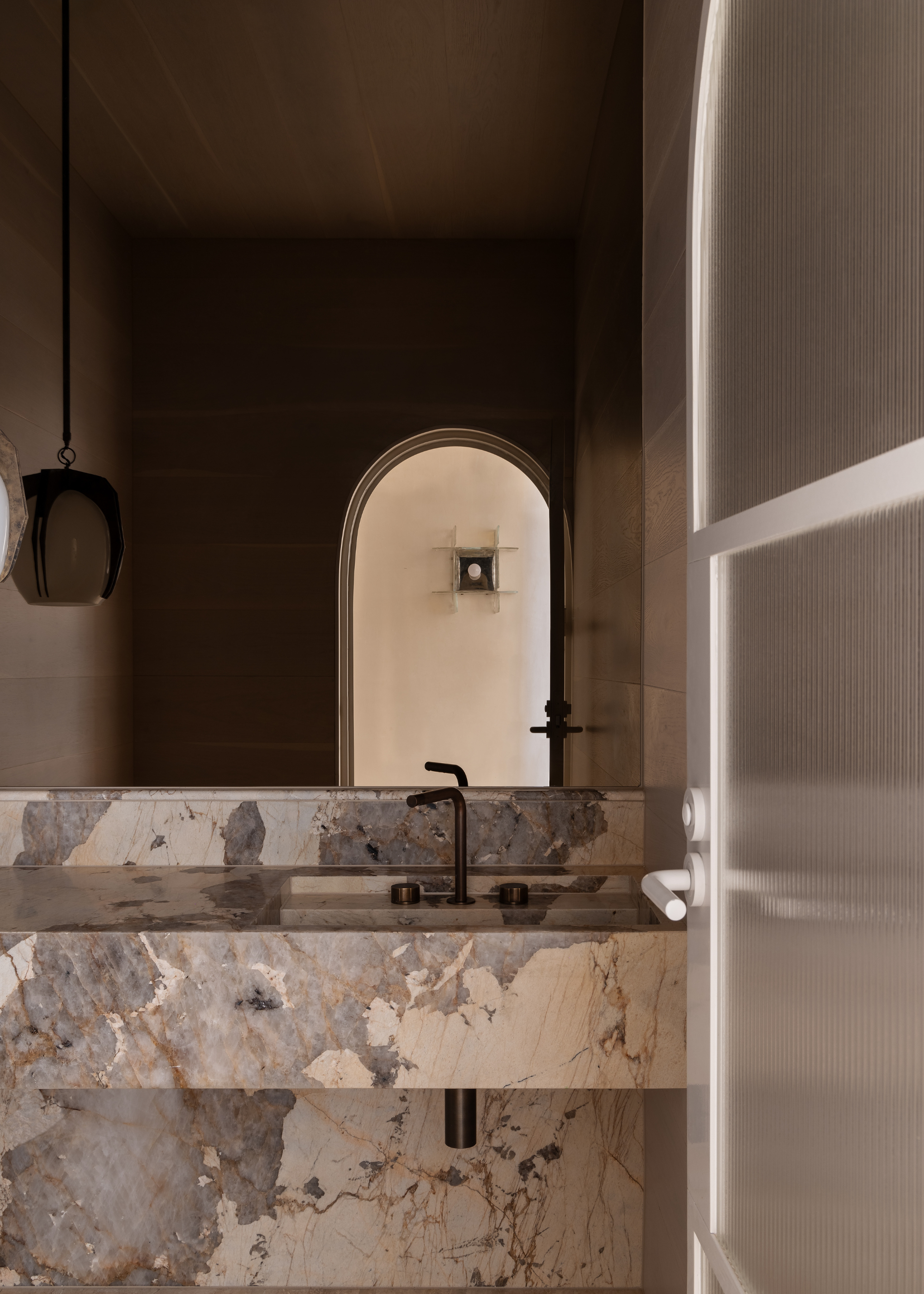 A minimalist bathroom with an arched doorway, a stone basin, a large mirror, and a hanging lamp