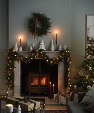 Christmas lights and a green garland hanging from a stone mantlepiece over a burning log fire. A green wreath hanging above in a sage green living room