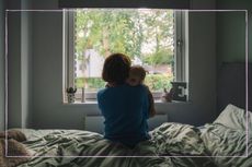 A woman pictured from behind looking out a window while cuddling a toddler
