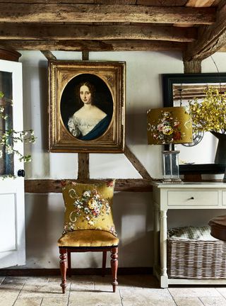 cottage hallway with open door, beams and gold floral cushion and matching lampshade and 18th-century style framed portrait of a woman and timbered walls