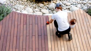 Top down shot of person applying stain halfway across timber decking with white stone in background