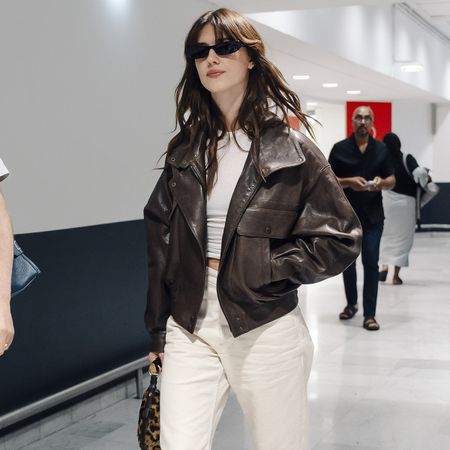 Daisy wears a brown leather jacket, white tee, and white jeans.