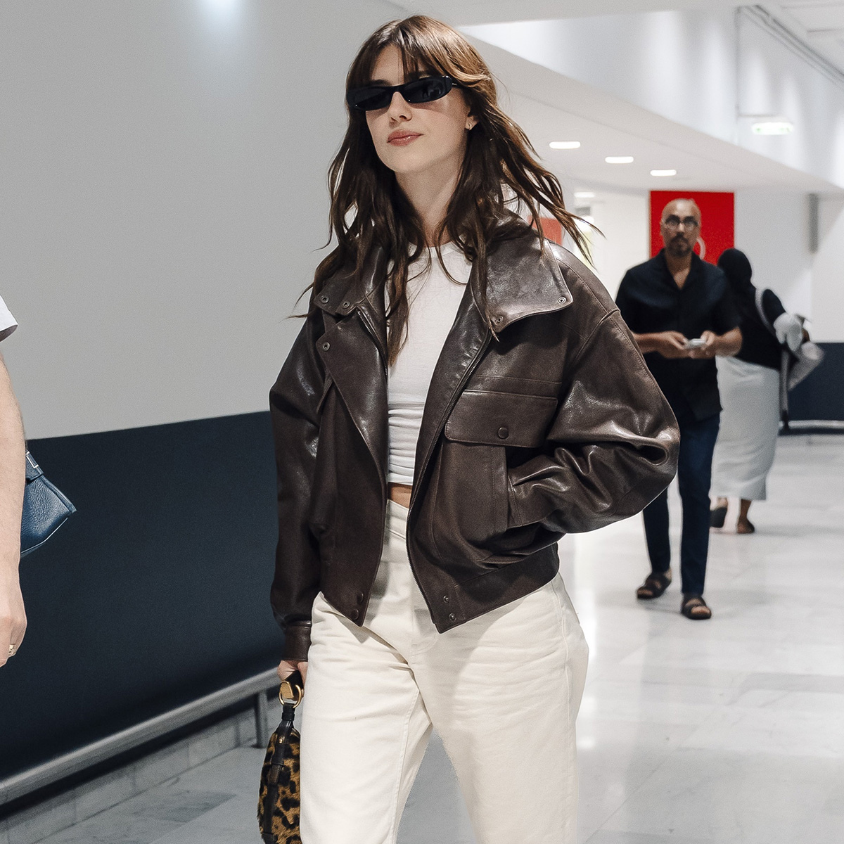 Daisy wears a brown leather jacket, white tee, and white jeans.