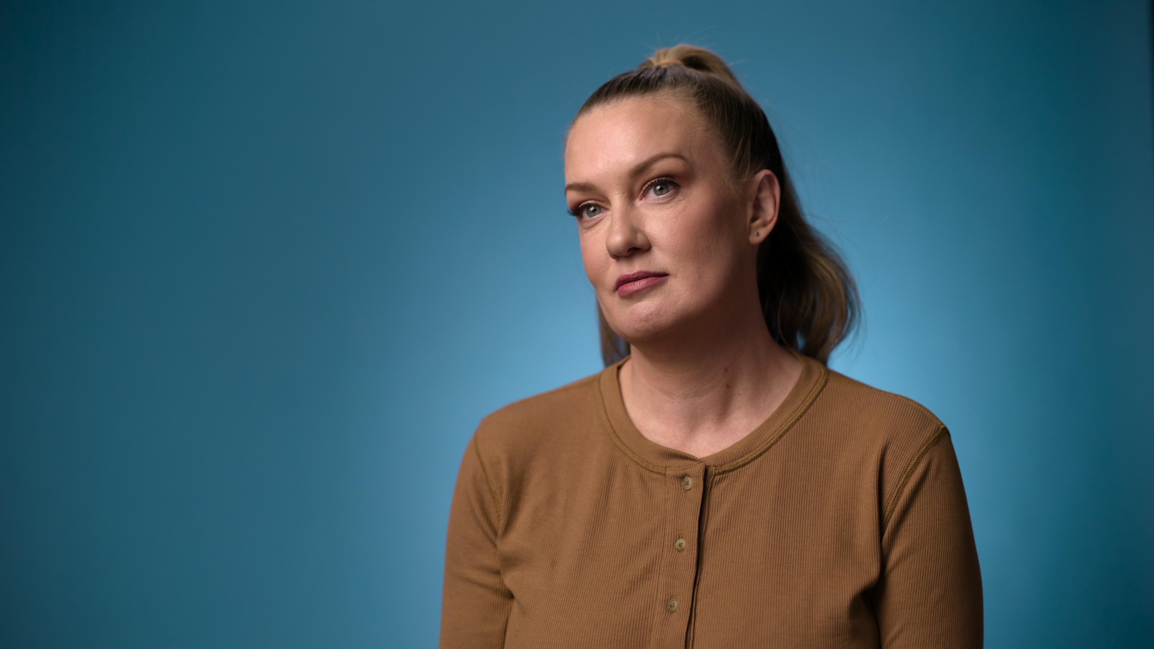 Former model and content-creator Joanie Sprague wears a brown cardigan while sitting in front of a blue backdrop, in the Netflix docuseries 'Reality Check: Inside America's Next Top Model.'