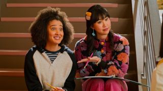 From left to right: Lee Rodriguez smiling sitting next to Ramona Young who is pointing at her while they sit on the stairs in Never Have I Ever.