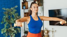 woman in a blue vest and orange bottoms raising a small yellow dumbbell in each hand to shoulder height. shot waist up, facing the camera with part of a blue wall and plant behind her 