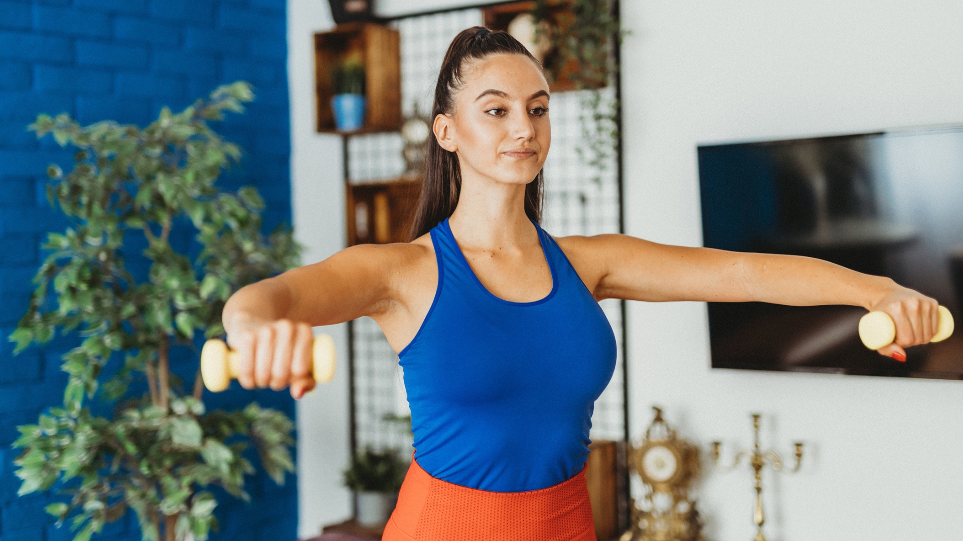 woman in a blue vest and orange bottoms raising a small yellow dumbbell in each hand to shoulder height. shot waist up, facing the camera with part of a blue wall and plant behind her