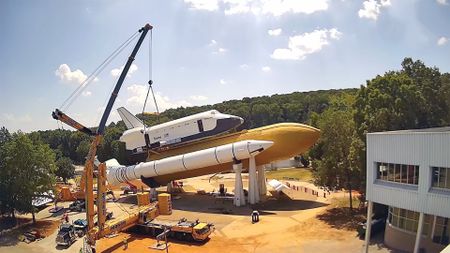 a crane lifts a full-size model of a nasa space shuttle onto its mock orange fuel tank and white solid rocket boosters in a horizontal position, with trees in the background