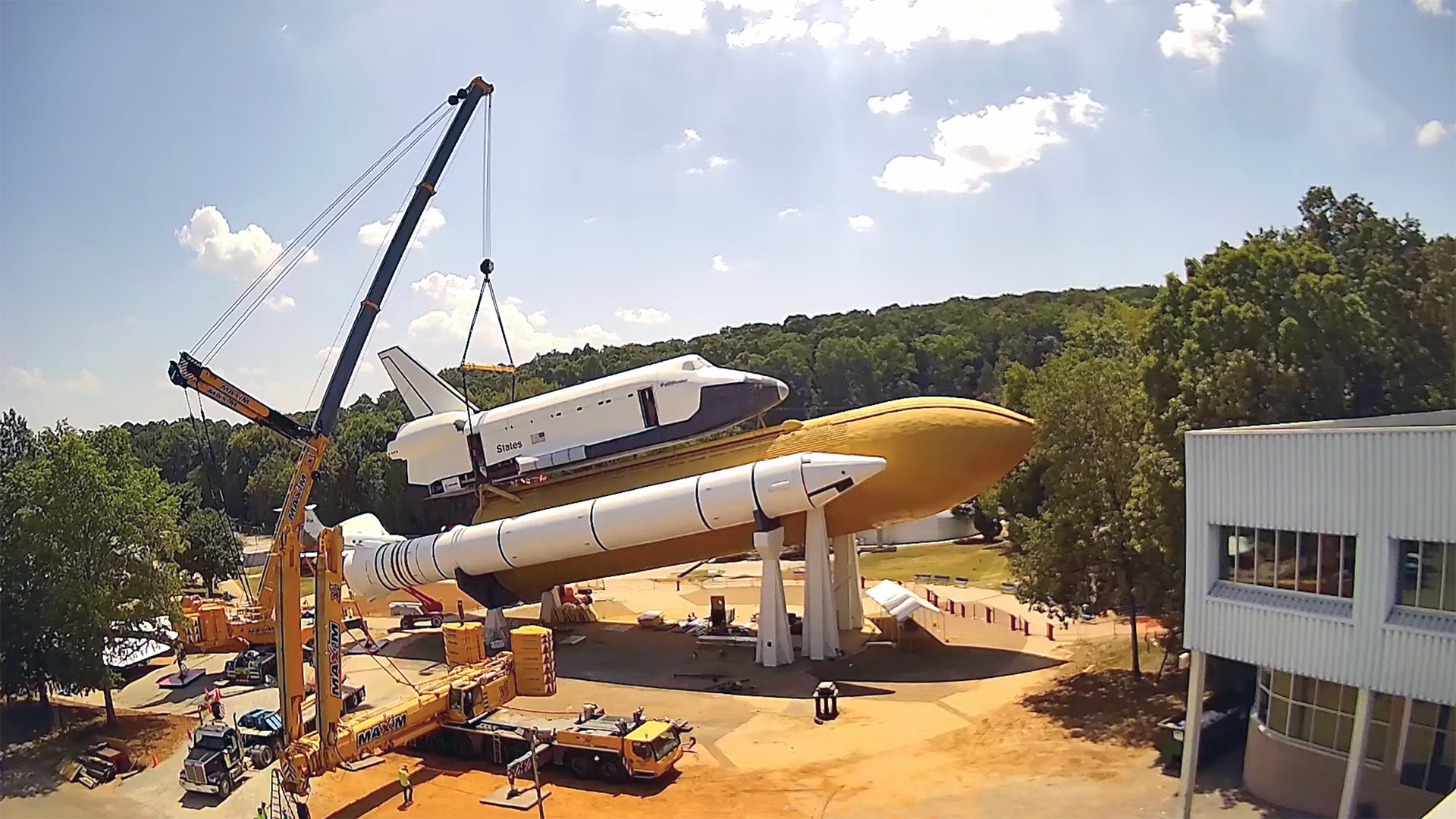 NASA Space Technology a crane lifts a full-size model of a nasa space shuttle onto its mock orange fuel tank and white solid rocket boosters in a horizontal position, with trees in the background