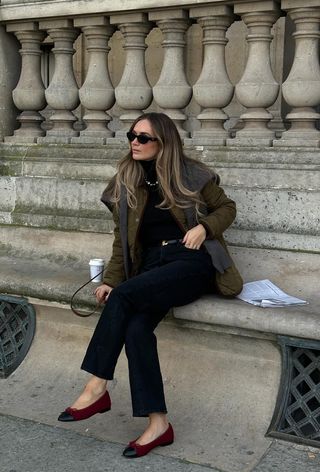 A black turtleneck outfit is shown in a photo of a woman sitting on a park bench wearing a green quilted jacket with a gray cardigan layered over it styled with a black turtleneck, black belt, skinny jeans, and red ballet flats