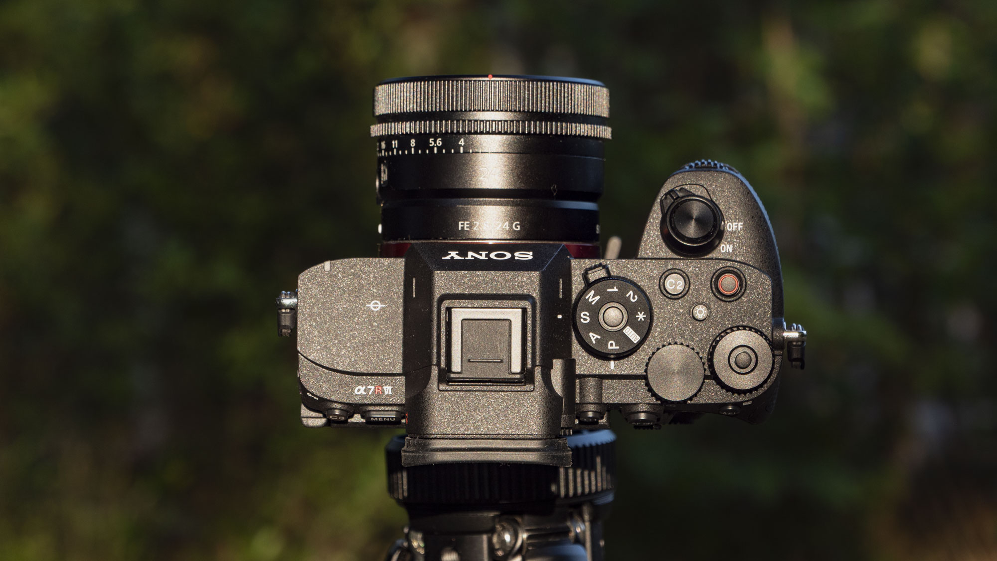 The Sony A7R VI camera with 24mm f/2.8 lens attached, it's mounted to a tripod outdoors and pointing up revealing the top plate, there's a natural background