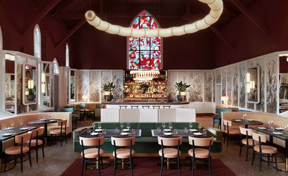 Interior view of Claudine restaurant, Singapore in a refurbished chapel. There is a dark wine coloured ceiling, an arched stained glass window, curved paper lantern lighting suspended from the ceiling, wall panels, green and light coloured seating, tables with tableware and a view of the bar
