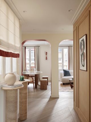 A hallway with neutral walls, console table and Roman blinds with a deep red fringed trim, leading into a living room with curtains lined with deep red edge