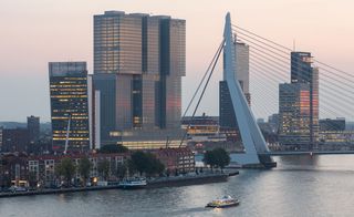 Exterior image of De Rotterdam's dramatic complex of three 150-meter-high towers, surrounding water, bridge, boat on the water, buildings, trees, dusk sky