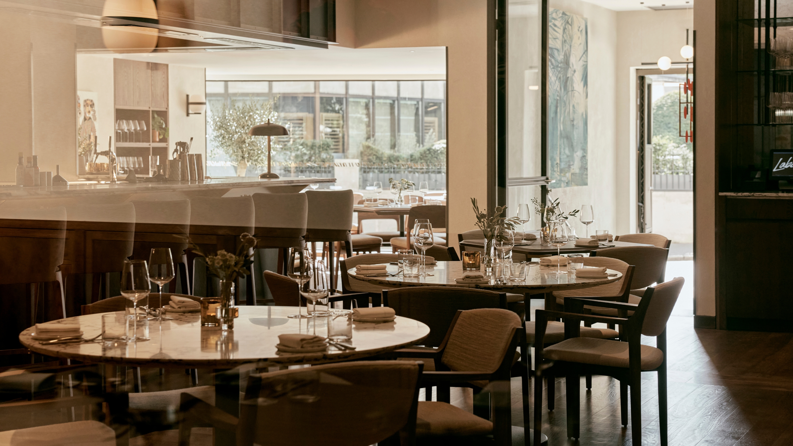 A naturally lit dining room with beautiful, elegant furnishings in textile and wood, striking artwork, and reflective table tops is photographed from behind a glass.